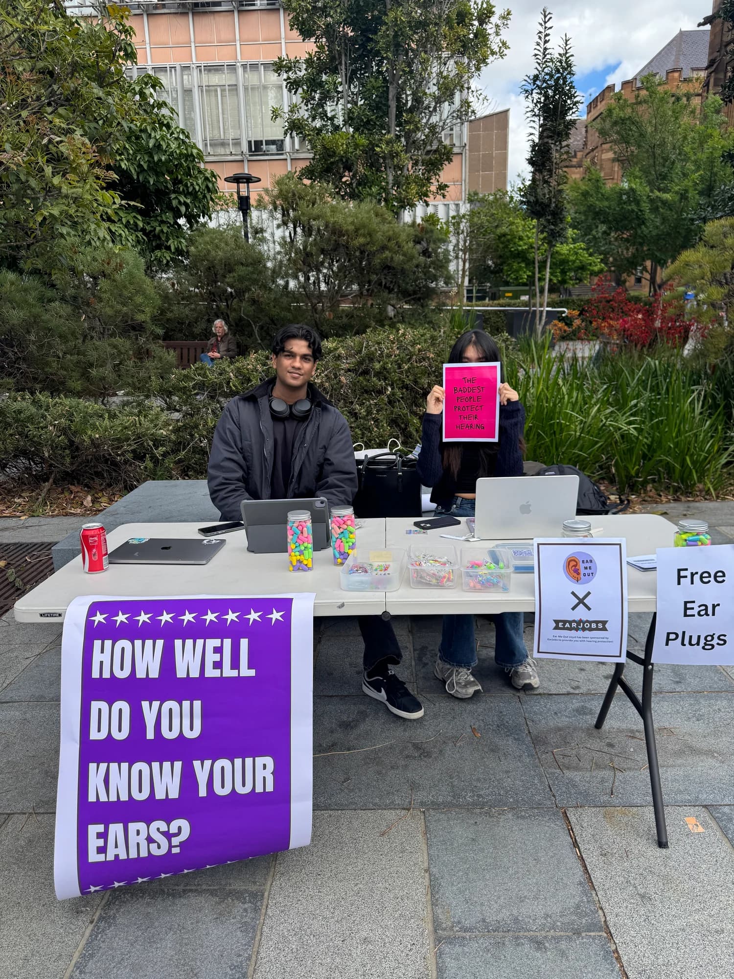 Two people behind the EarMeOut campus stall with jars of colourful earplugs, keychains, and hearing-awareness posters.